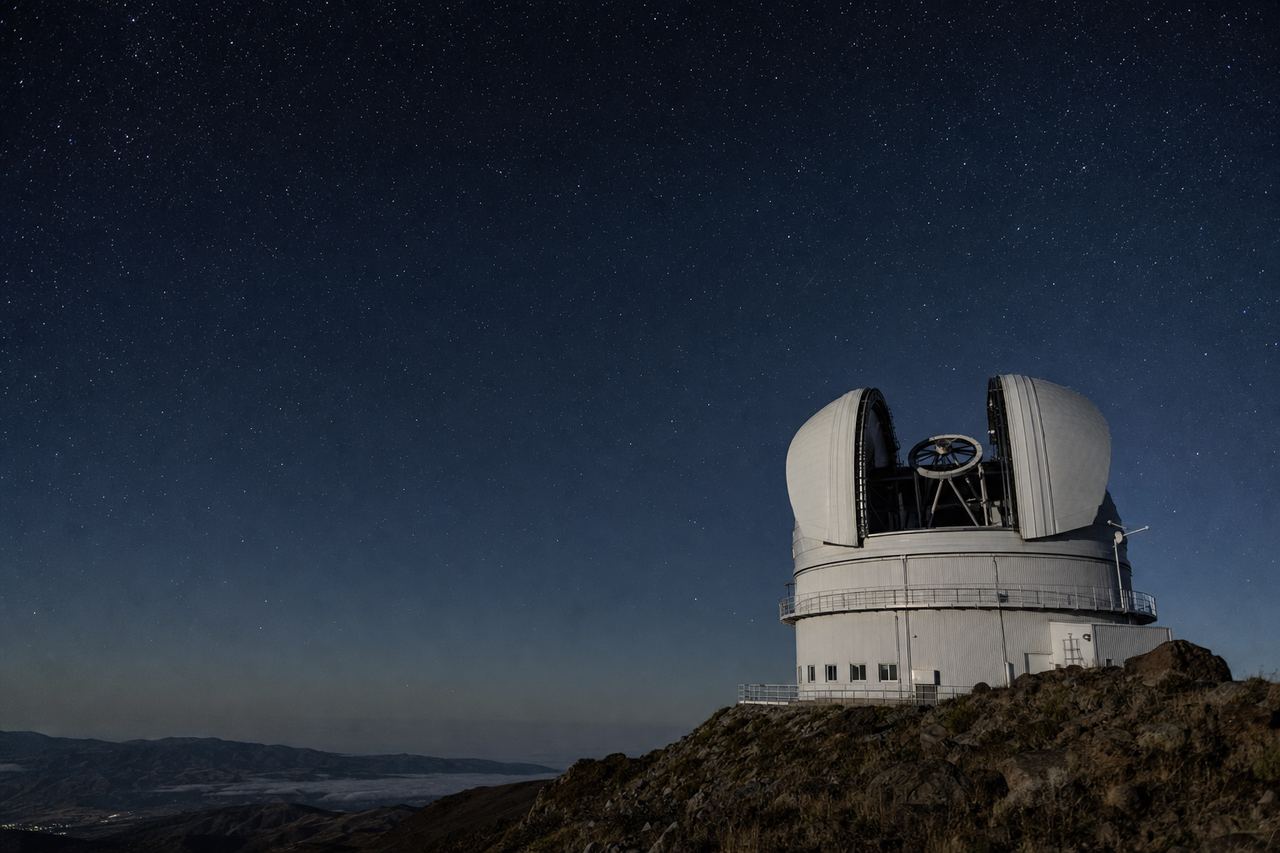 A mountaintop astronomical observatory at night, its dome open to a sky full of stars above a dark ridgeline.